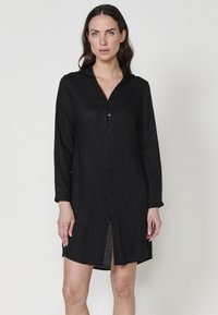 Rochie neagră cu mâneci lungi, dintr-un material ușor. Are un închidere cu nasturi în față, guler și fante laterale pentru ventilație.