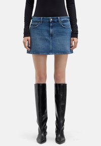 Jeans-Minirock in Blau mit klassischem Fünf-Taschen-Design, einem Bund mit Gürtelschlaufen und einem geraden Saum, kombiniert mit schwarzen Overknee-Lederstiefeln.