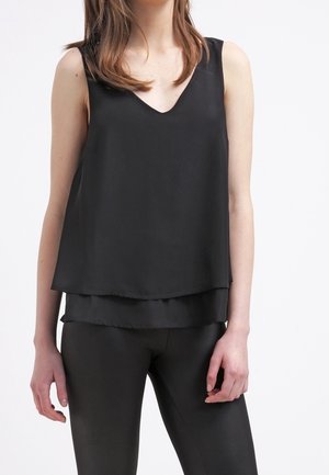 Femme portant un haut noir sans manches à col en V superposé et un pantalon noir ajusté, debout devant un fond blanc uni.