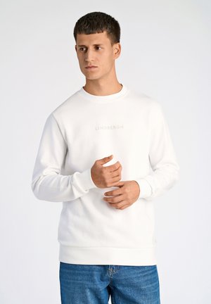 Sudadera blanca con cuello redondo y mangas largas. Presenta "LINDBERGH" impreso en una fuente sutil en el pecho. Combinada con vaqueros azules.