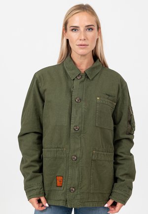 Blonde vrouw draagt een olijfgroene jas met knopen, met meerdere voorzakken en een klein bruin label op de onderste linkerzak.