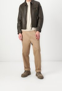 Veste en cuir brun avec poches zippées, pull beige, pantalon beige et bottes à lacets brunes. Design simple avec une coupe décontractée.