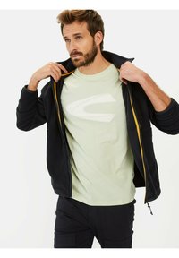 Marinegrøn fleecejakke med gule lynlåse, lagd over en lys grøn t-shirt med et hvidt logo, båret af en mand med lyst hår.