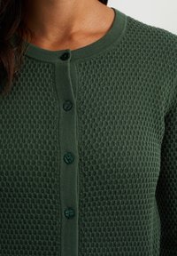 Grön strukturerad kofta med ett bikakestruktur, rund halsringning och sex mörkgrön knappar framtill.