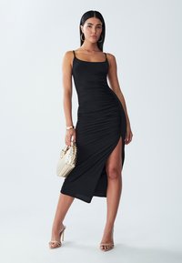 Black fitted dress with spaghetti straps, ruching detail, and a thigh-high slit. Paired with a textured beige handbag and strappy heels.