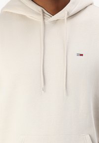 Sudadera de color crema hecha de una tela suave, con un bolsillo canguro en la parte delantera, capucha ajustable con cordones y un pequeño logotipo bordado en el pecho.