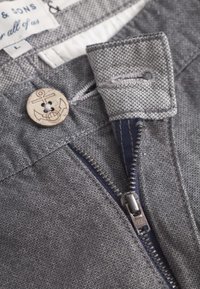Tessuto grigio con una trama texturizzata, caratterizzato da un bottone di legno con design a ancora e una cerniera metallica di un blu a contrasto.