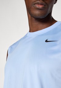 Jasnoniebieska bezrękawnik sportowy wykonany z gładkiego materiału, z czarnym logo Nike na górnej lewej stronie. Elastyczny krój i okrągły dekolt.