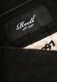 Etiqueta de tela negra con el nombre de la marca "Reell" en escritura blanca y "est. 1997" en una fuente más simple sobre un fondo beige.