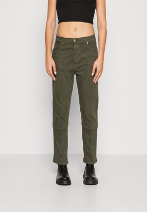 Mujer que lleva pantalones verde oliva con bolsillos delanteros tipo parche, top corto negro y botas negras hasta el tobillo, de pie contra un fondo claro y liso.