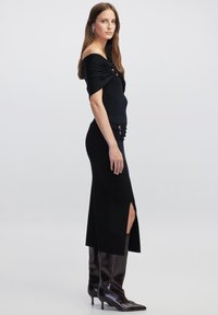 Černé off-shoulder bodycon šaty s ribbed texturou, mají rozparek na boku a nařasené detaily v pase, v kombinaci s hnědými kozačkami na vysokém podpatku.