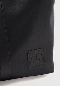 Μαύρη δερμάτινη επιφάνεια με ανάγλυφο λογότυπο Armani Exchange που περιλαμβάνει τα γράμματα A και X μέσα σε τετράγωνο έμβλημα.