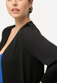 Svart ribbad cardigan med lång ärm och öppen design, bärs över ett blått plagg, med en slät textur. Gyllene hoop-örhängen synliga.