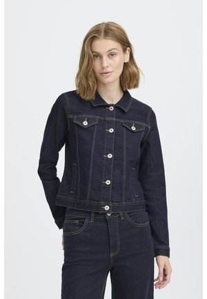 Femme aux cheveux châtain clair mi-longs, portant une veste en jean bleu foncé et un jean assorti, sur un fond clair uni.