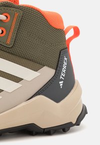 Lehká turistická bota s olivově zeleným textilním svrškem, béžovou mezipodešví, černým podpatkem a jasně oranžovými akcenty. Logo viditelné na boku.