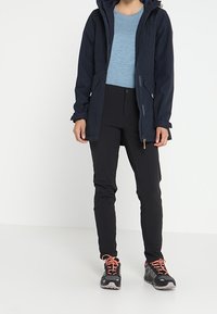 Marinblå vattentät jacka med huva, i kombination med en ljusblå topp och svarta byxor. Skorna har orangea detaljer och en texturerad sula.