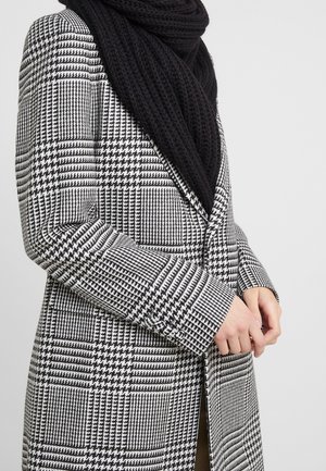 Femme portant un manteau à motif pied-de-poule noir et blanc ainsi qu'une épaisse écharpe noire, les mains jointes devant un fond uni.