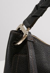 Gros plan d’un sac à main en cuir noir texturé avec une bandoulière tressée attachée par une boucle en métal argenté et des détails de couture marron.