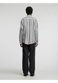 Svart-hvitt stripete skjorte med lange ermer, løs passform, buet nederkant og kontrasterende mørke bukser. Stoffet virker glatt og lett.