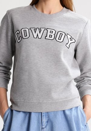 Kvinde iført en grå sweatshirt med "COWBOY" trykt på brystet, kombineret med lyseblå jeans, hænderne i lommerne.
