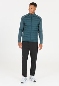 Veste rembourrée en teal avec fermeture éclair, manches lisses, pantalon noir et chaussures de sport blanches. La veste présente un extérieur texturé et un design ajusté.