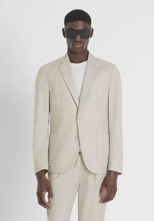 Veste de costume beige ajustée avec deux poches avant, fermeture à un bouton, texture lisse, associée à une chemise en tricot côtelé blanche et des lunettes de soleil noires.