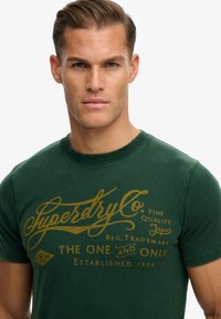 Mørkegrøn kortærmet t-shirt lavet af bomuld, med gyldne bogstaver og et vintage logo design på forsiden.