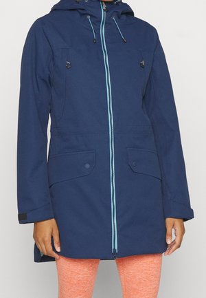 Person wearing a navy blue hooded jacket with light blue zipper and orange leggings, standing against a plain background.