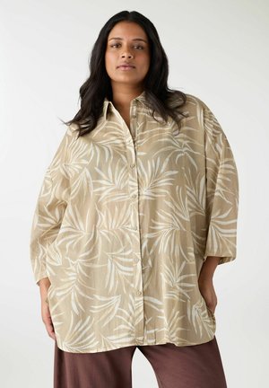 Donna con capelli lunghi e scuri che indossa una camicia beige oversize con bottoni e motivo a foglie bianche, e pantaloni marroni, in piedi davanti a uno sfondo semplice.