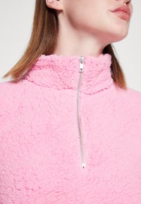 Pullover in pile rosa con colletto alto e chiusura con zip argentata. La texture è morbida e soffice, caratterizzata da un colore uniforme senza motivi.