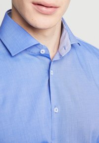 Muž v modré košili s knoflíky a pruhovaným vnitřním límcem, s viditelnými jen rty a bradou.