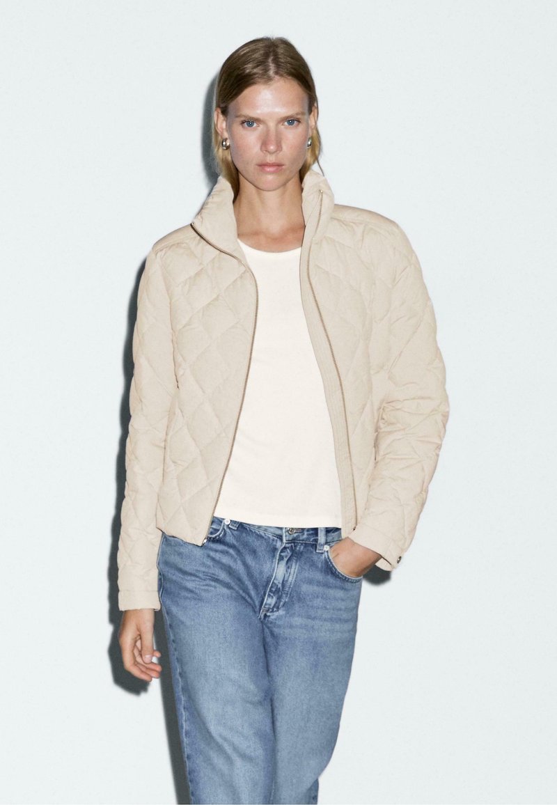 Beige gewatteerde jas met een hoge kraag en rits, gedragen over een witte top en lichtblauwe jeans, tegen een neutrale achtergrond.