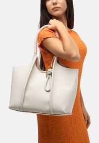 Sac fourre-tout en cuir blanc avec des poignées tressées et un détail de nœud décoratif. Texture lisse, design spacieux, sans matériel visible.
