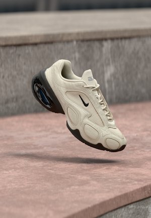 Baskets Nike beige clair avec semelle noire et coussin d'air, en plein vol au-dessus des marches en pierre dans un environnement extérieur.