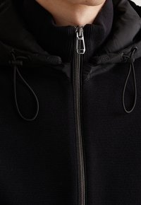 Gros plan d'une veste en maille noire avec une fermeture éclair argentée, un col montant et des cordons de capuche réglables de chaque côté.