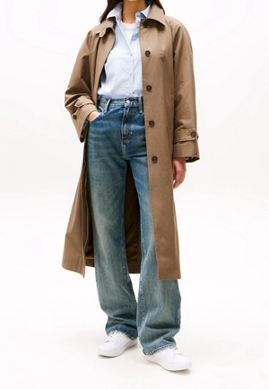 Kvinde iført beige trenchcoat over lyseblå skjorte, blå jeans og hvide sneakers, stående med den ene hånd i lommen.