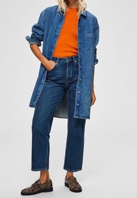 Jeansskjorta, orange ribbad tröja och mörkblå högmidjade jeans. Texturerade svarta loafers med gulddetaljer. Avslappnad lager-på-lager-outfit.