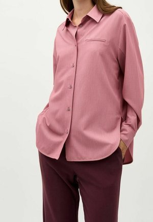 Kobieta ubrana w luźną, długorękawową, przygaszonoróżową koszulę na guziki z kieszenią na piersi z rozcięciem, w połączeniu z ciemnoczerwonymi spodniami, z rękami w kieszeniach.