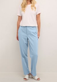 Pantaloni su misura blu con una texture liscia e pieghe frontali, abbinati a una maglietta bianca semplice e scarpe aperte bianche.