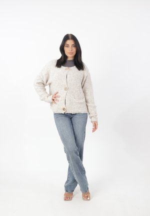 Cardigan beige chiaro e morbido con scollatura tonda, tre bottoni in legno e due tasche anteriori. Indossato sopra una maglietta grigia e jeans.