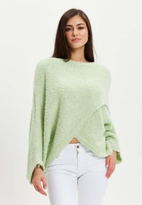Light green, soft, textured pullover with a loose, asymmetrical design and wide sleeves, paired with white pants.