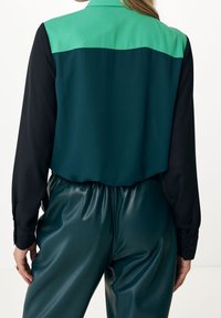 Button-up shirt with a green top panel and black sleeves; paired with shiny, dark green pants. Smooth texture, modern design.