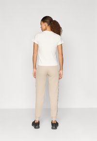 Ανοιχτό μπεζ T-shirt με κοντά μανίκια, συνδυασμένο με μπεζ φόρμες που διαθέτουν πλευρικές ρίγες. Το σύνολο ολοκληρώνεται με μαύρα παπούτσια.