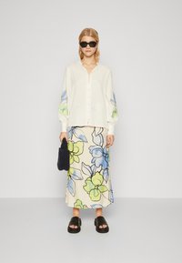 Witte knop-up blouse met kleurrijke bloem borduursels op de mouwen, gecombineerd met een crème rok met blauwe en groene bloemmotieven. Zwarte sandalen.