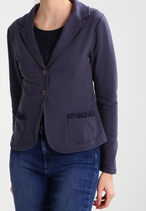 Vrouw draagt een getailleerd donkerblauw colbert met twee knopen en opgestikte zakken, gecombineerd met een blauwe spijkerbroek tegen een effen achtergrond.