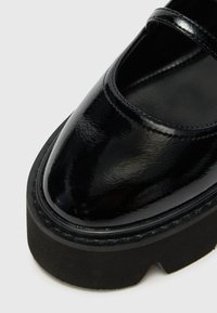 Black patent leather shoe with a rounded toe, featuring a two-strap design and thick black rubber sole, showcasing a glossy finish.