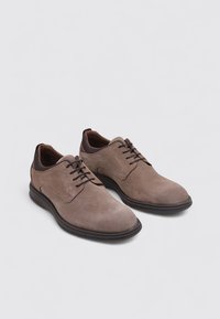 Zapatos de ante marrón con una textura suave, punteras redondeadas, costuras sutiles y suelas de goma negra. Presentan cordones y elementos de diseño mínimos.