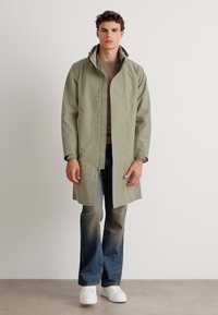 Chaqueta impermeable de color verde claro con capucha, cierres a presión en el frente y manga larga, superpuesta sobre una blusa marrón y combinada con jeans azules y zapatillas blancas.