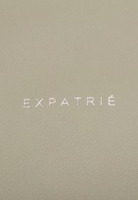 Surface texturée beige clair avec le mot « EXPATRIÉ » brodé en fil blanc au centre.