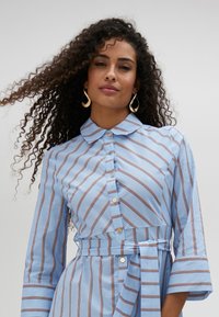 Light blue shirt dress with horizontal red and white stripes, buttoned front, collar, and tied belt at the waist; three-quarter sleeves.